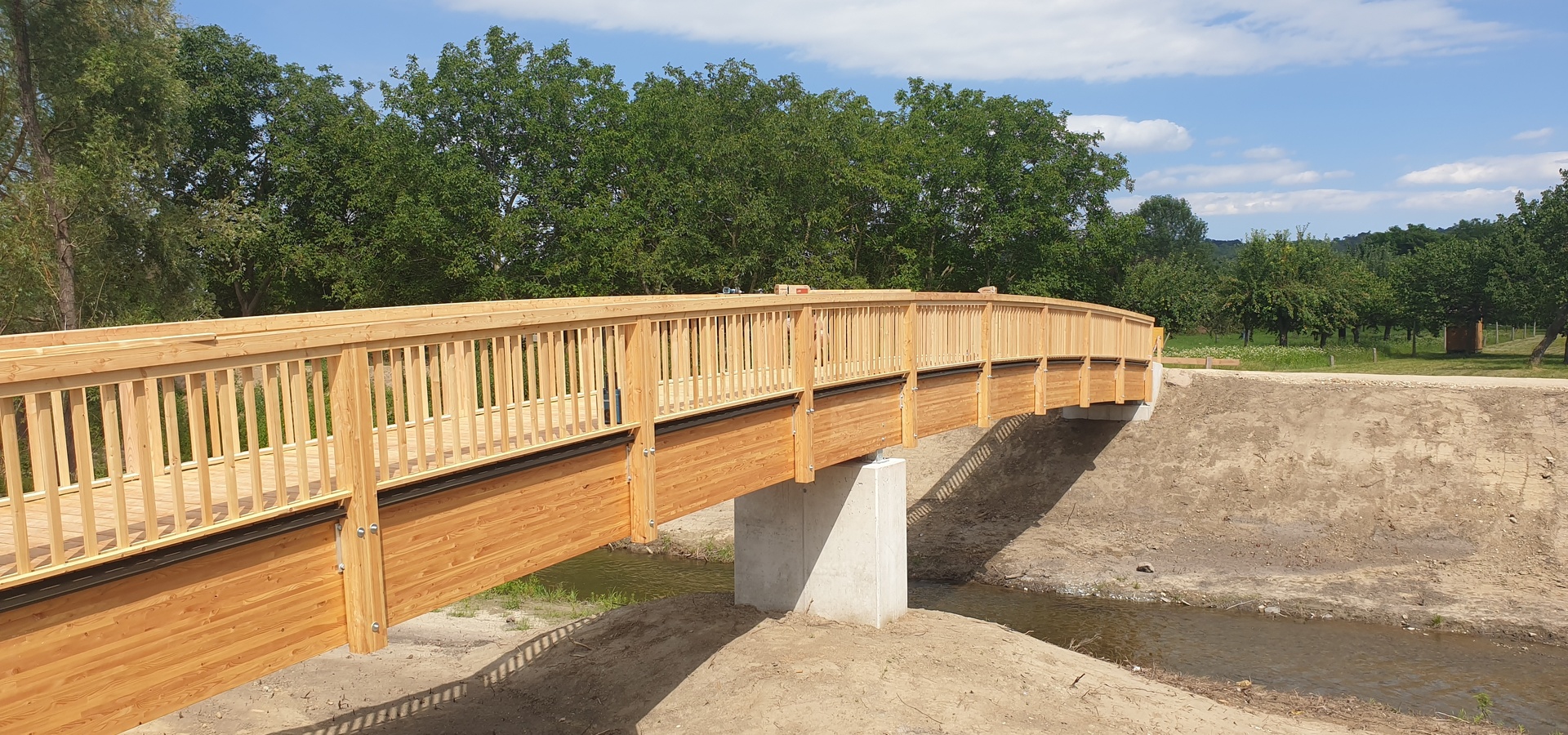 Eine hölzerne Fußgängerbrücke überspannt einen kleinen Bach, umgeben von Bäumen und einem klaren Himmel.