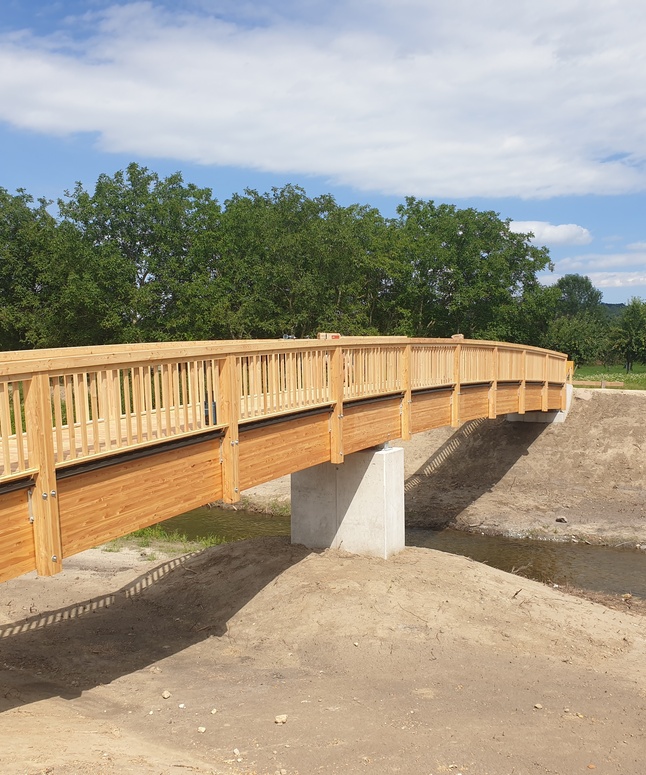 Eine hölzerne Fußgängerbrücke überspannt einen kleinen Bach, umgeben von Bäumen und einem klaren Himmel.
