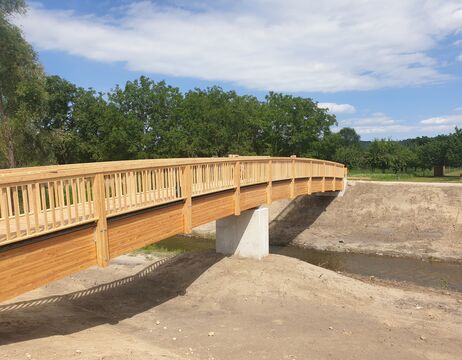 Eine hölzerne Fußgängerbrücke überspannt einen kleinen Bach, umgeben von Bäumen und einem klaren Himmel.