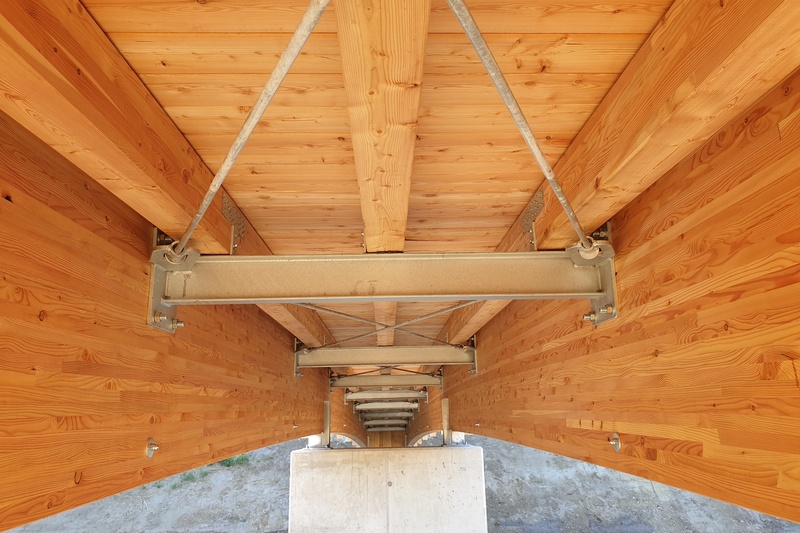 Blick von unten auf eine Holzbrücke mit Metallstützen und -trägern, die konstruktive Details zeigt.