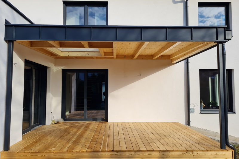 Moderner Innenhof mit Holzterrasse und einer schwarzen Pergola mit Metallrahmen, die an ein helles Haus angebaut ist.