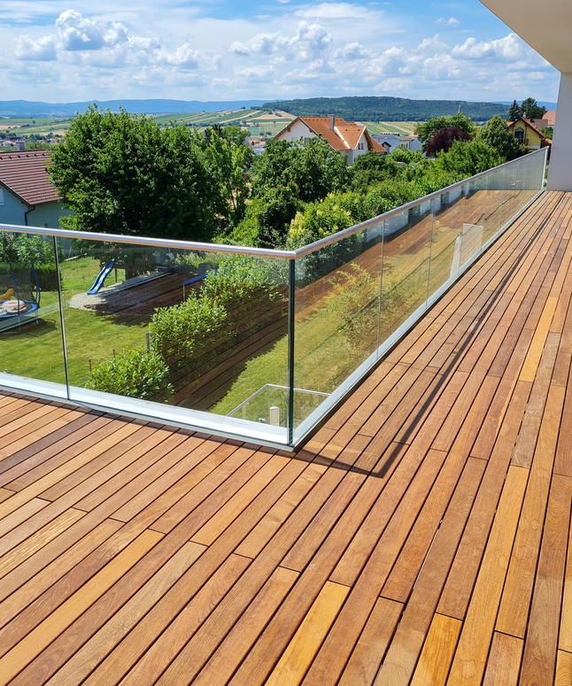 Der Holzbalkon mit Glasgeländer bietet an einem sonnigen Tag einen Blick auf einen grasbewachsenen Hinterhof und die Häuser der Nachbarschaft.