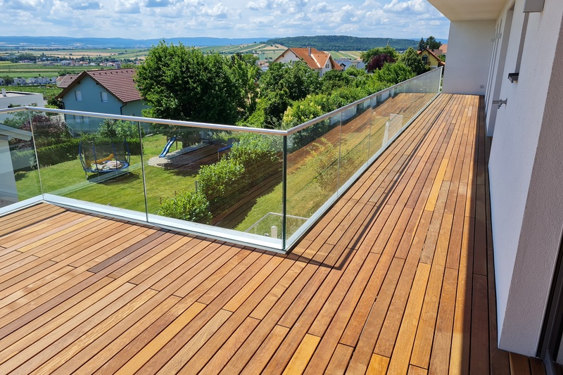Der Holzbalkon mit Glasgeländer bietet an einem sonnigen Tag einen Blick auf einen grasbewachsenen Hinterhof und die Häuser der Nachbarschaft.